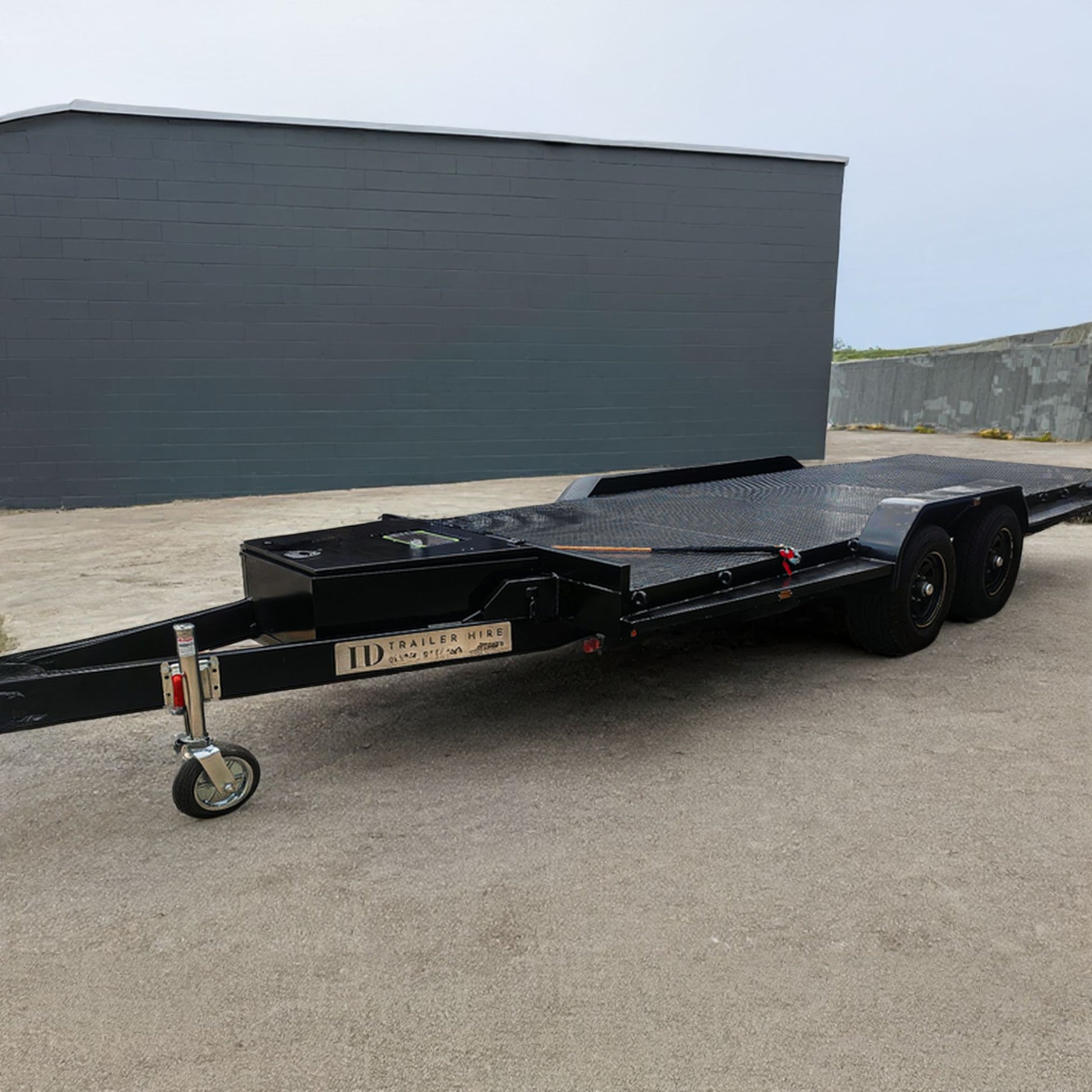 Trailer Hire Brisbane - Black flatbed trailer parked on a concrete surface with a dark building in the background.