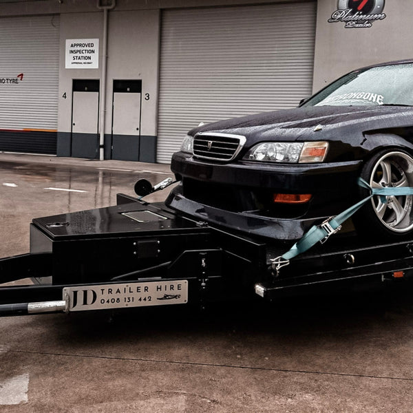 Black car being towed by a car trailer hire with a towing company's name on it, in front of a garage.