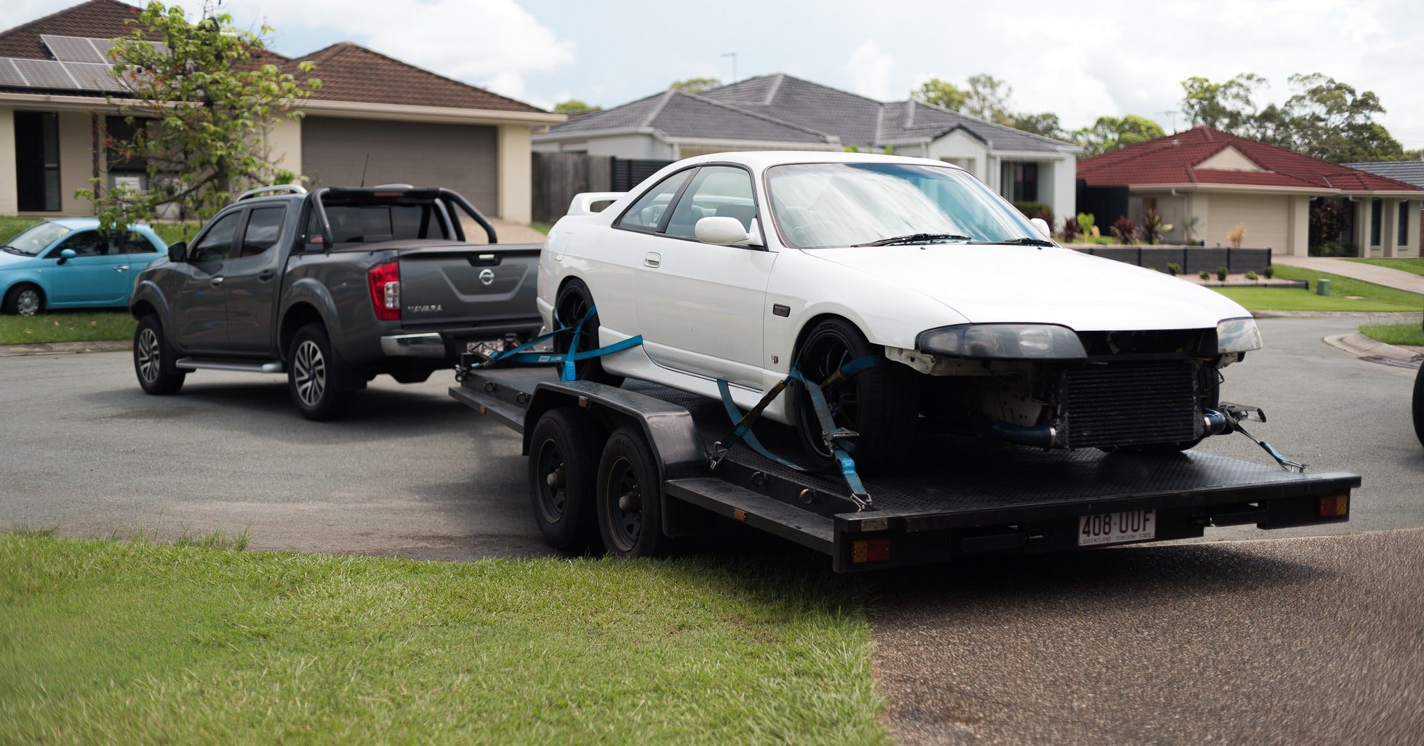 White car on a car trailer hire being towed by a black pickup truck in a residential area.
