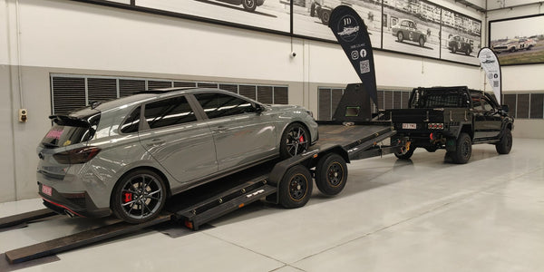 Gray car on a car trailer hire inside a garage with a black truck in the background.